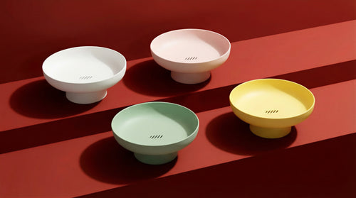 Runda Bowl by Maxim Vogel shown in Chalk, Blush, Sage and Citron, a colourful collection of sculptural matte fruit bowls on a deep red surface.