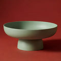 Runda Bowl in Sage by Maxim Vogel, a sculptural matte fruit bowl photographed against a deep red background.